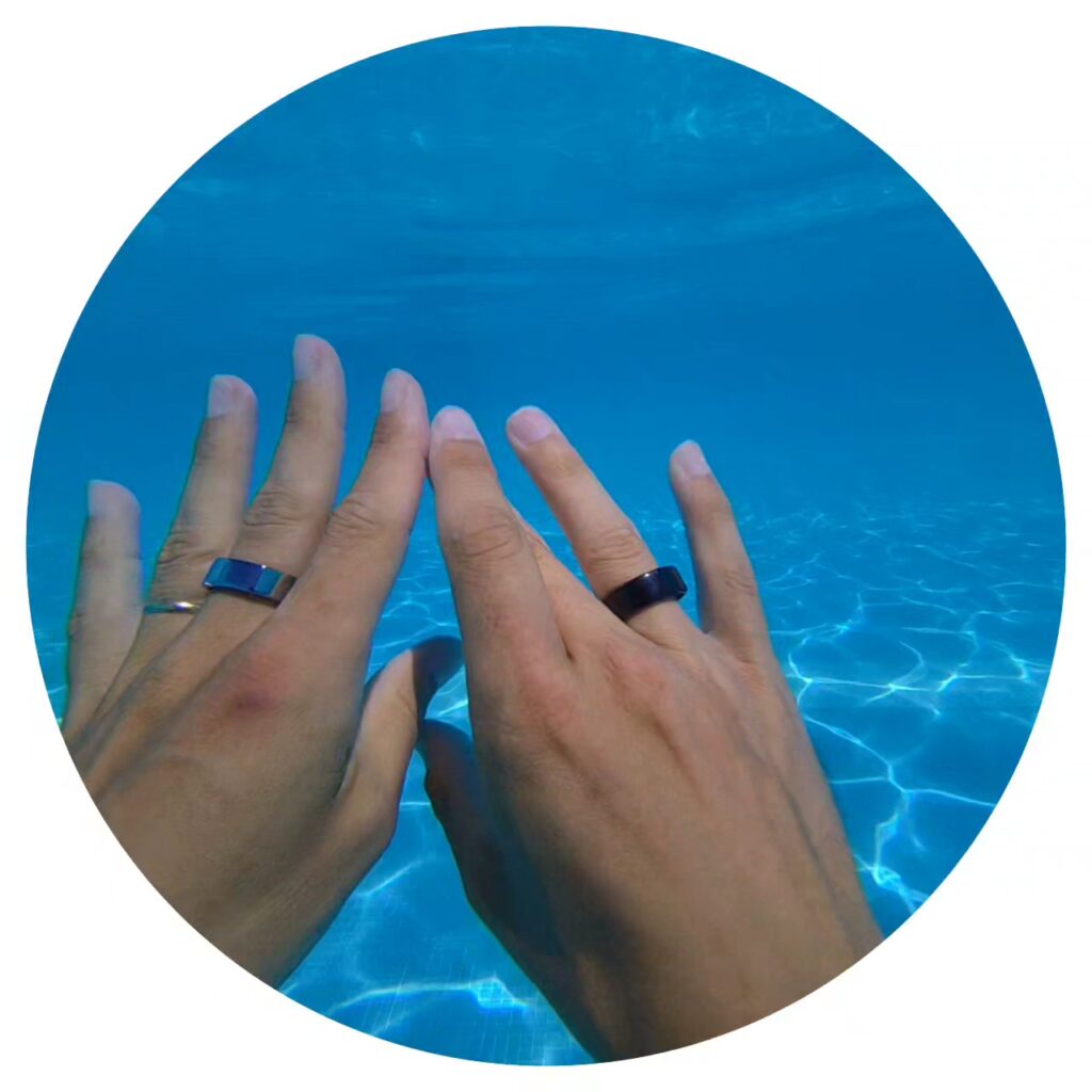 Two hands wearing Xiaomi Smart Ring and Oura Ring submerged underwater in a clear blue swimming pool, demonstrating their waterproof features in a real-life setting