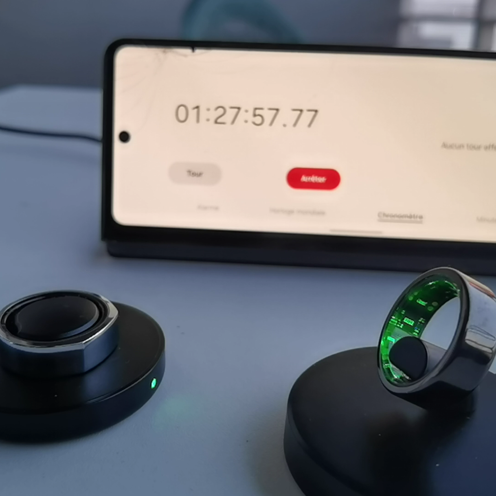 Xiaomi Smart Ring and Oura Ring placed on their chargers next to a smartphone displaying a timer at 1 hour, 27 minutes, and 57 seconds, indicating the duration of charging time for both devices